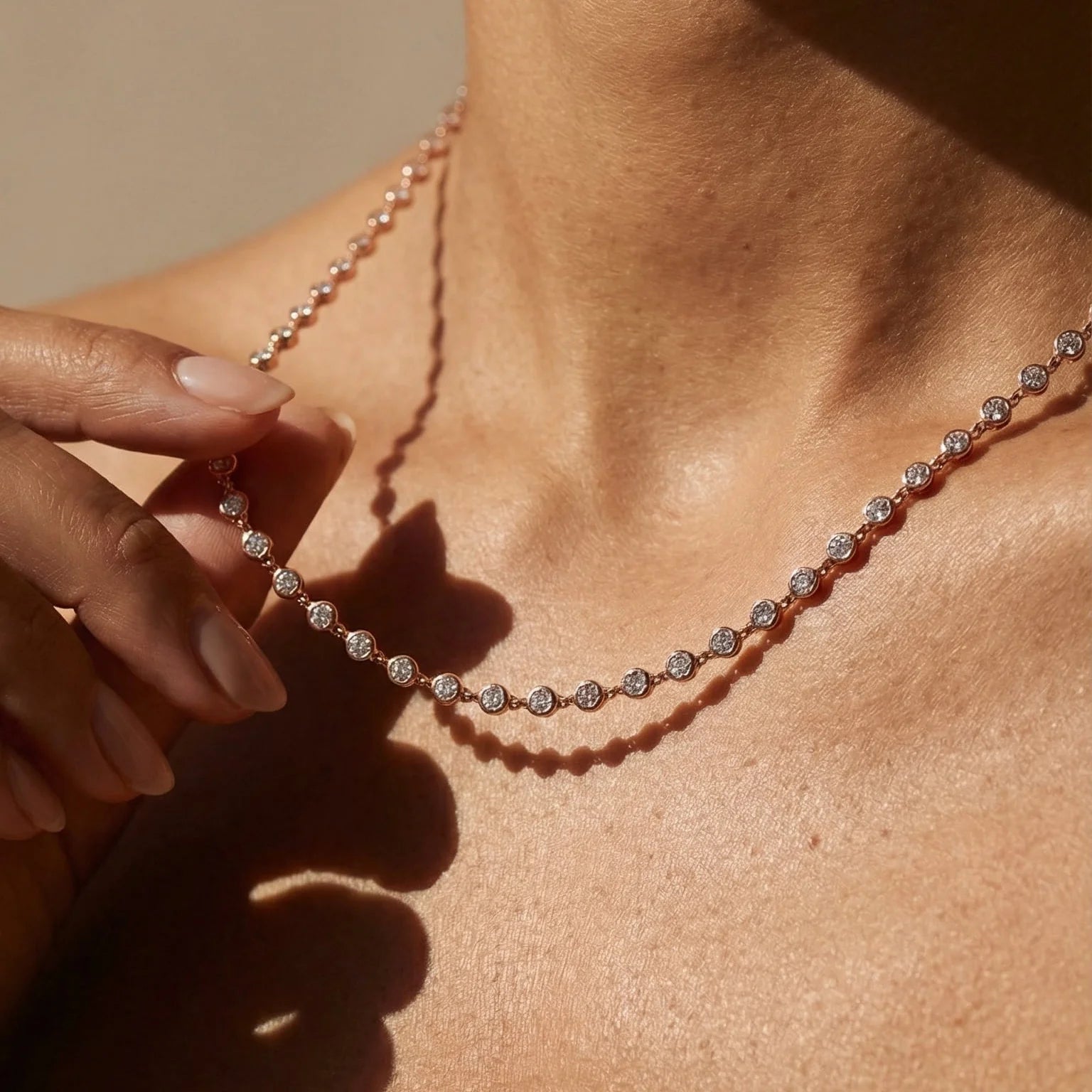 A close-up lifestyle shot of a delicate tennis necklace with round-cut clear stones set in a rose gold or pinkish silver-toned metal, worn on a model's neck with a hand gently touching one of the front stones.