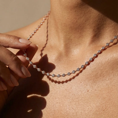A close-up lifestyle shot of a delicate tennis necklace with round-cut clear stones set in a rose gold or pinkish silver-toned metal, worn on a model's neck with a hand gently touching one of the front stones.