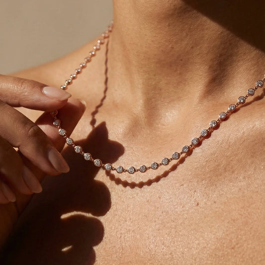 A close-up lifestyle shot of a delicate tennis necklace with round-cut clear stones set in a rose gold or pinkish silver-toned metal, worn on a model's neck with a hand gently touching one of the front stones.