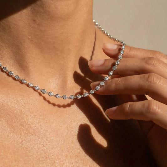 A close-up of a delicate silver tennis necklace with small round-cut stones worn on a model's neck, with a hand gently touching the chain.