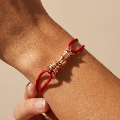 A close-up of a person's wrist wearing a red cord bracelet with a rose gold chess motif, set against a solid beige background.