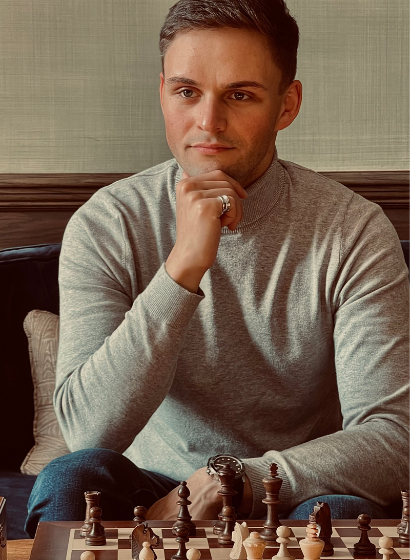 Man in a gray sweater sitting at a chessboard with a thoughtful expression.
