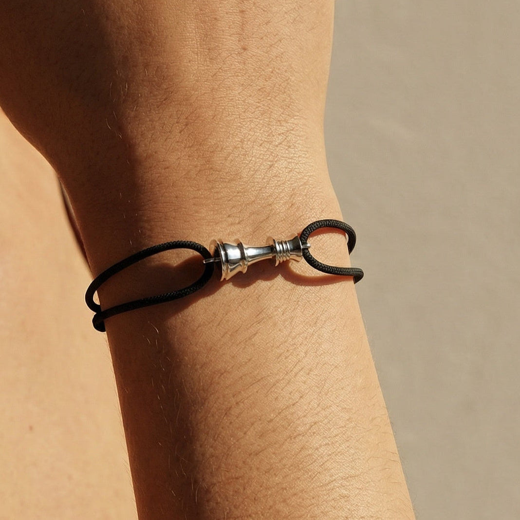 Close-up of a hand wearing a black cord  bracelet with white gold chess queen charm on a neutral background