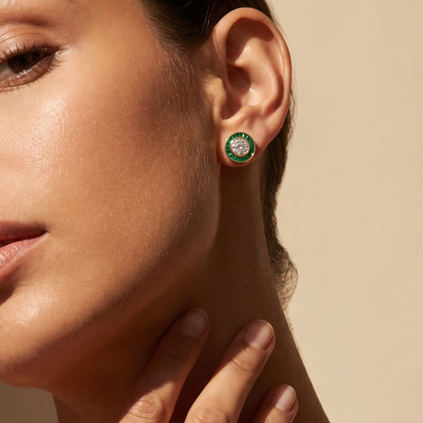 Close-up of a woman wearing a round stud earring with a green outer ring and a gold inner setting holding small clear stones, shown against a warm beige background.