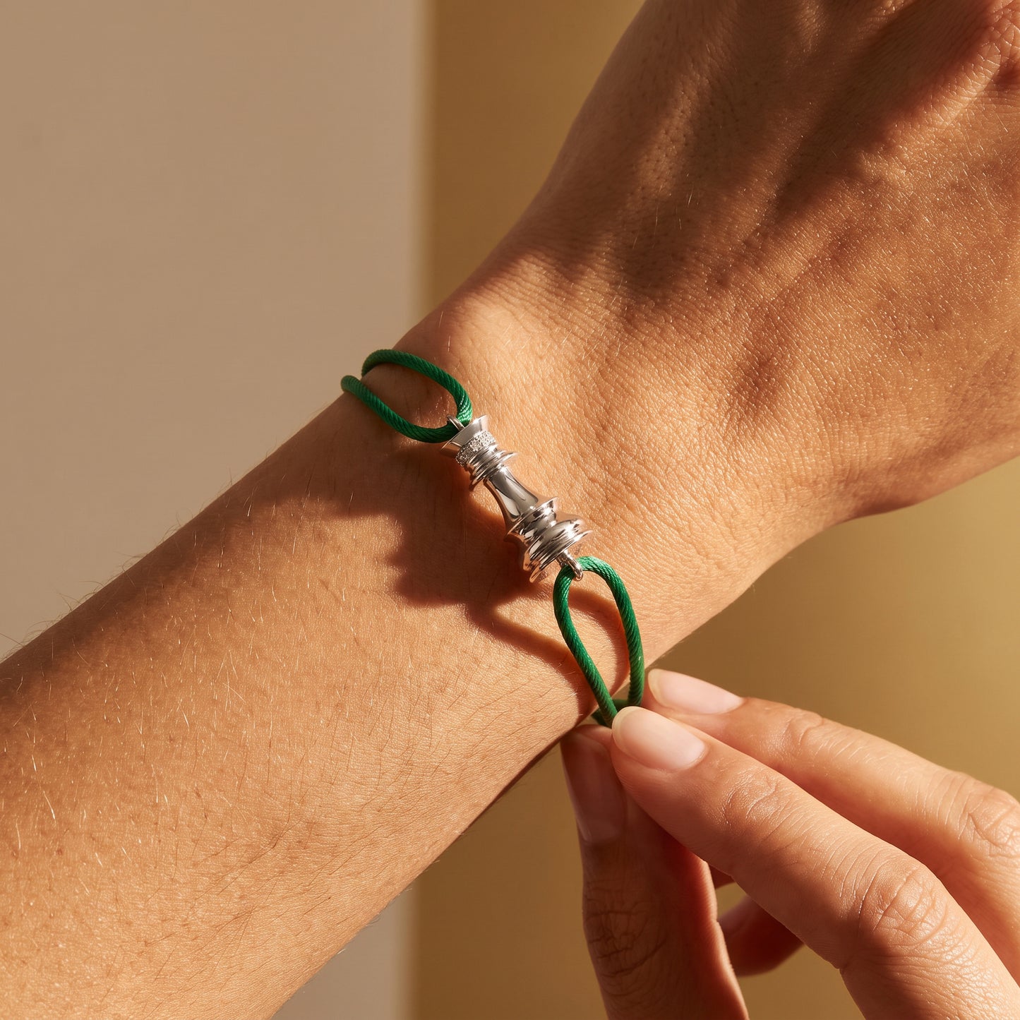 A close-up of a person's wrist wearing a green cord bracelet with a white gold chess motif, with another hand adjusting the clasp against a warm beige background.