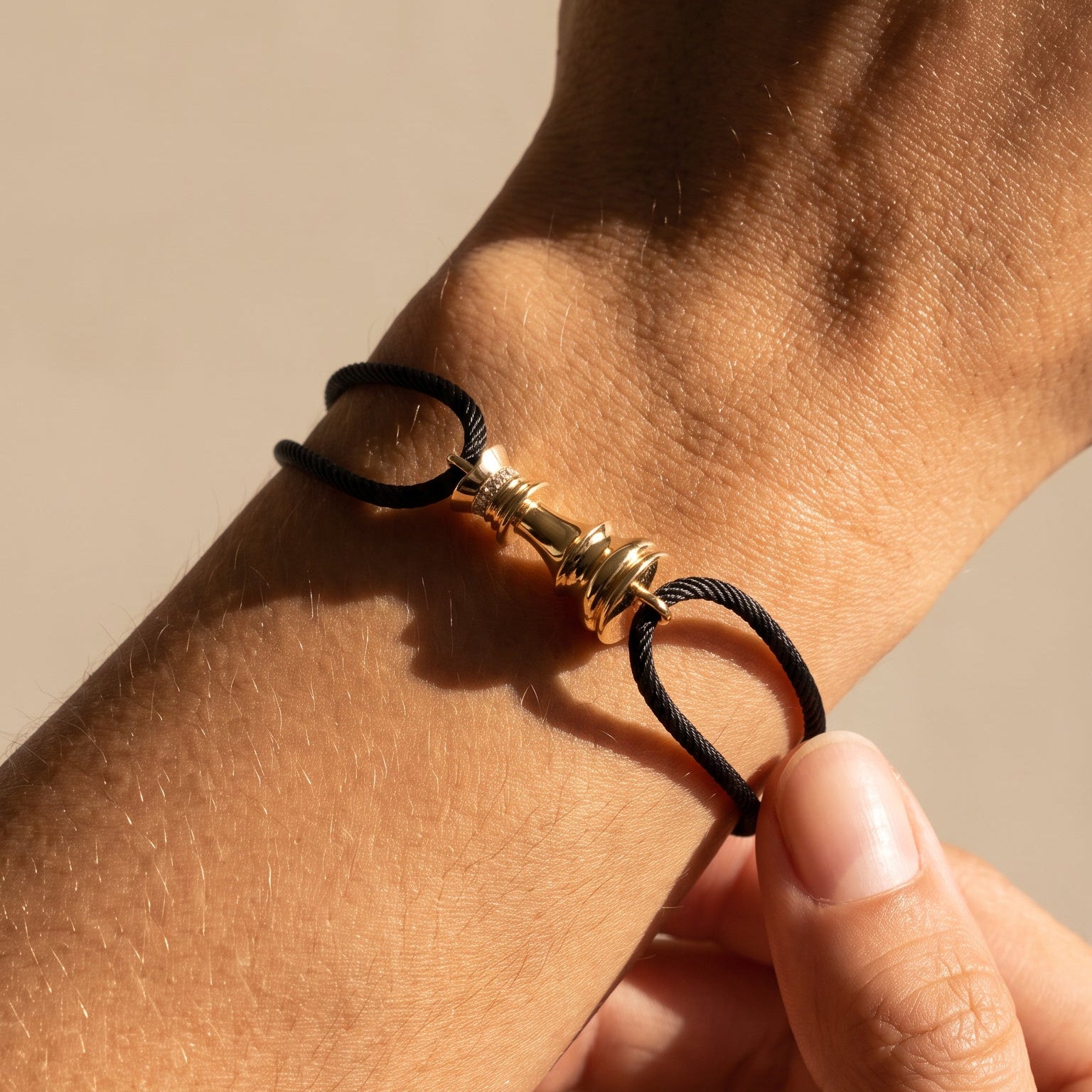 A close-up of a black cord bracelet with a gold-toned chess motif, worn on a person's wrist with another hand adjusting it.