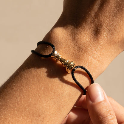 A close-up of a black cord bracelet with a gold-toned chess motif, worn on a person's wrist with another hand adjusting it.