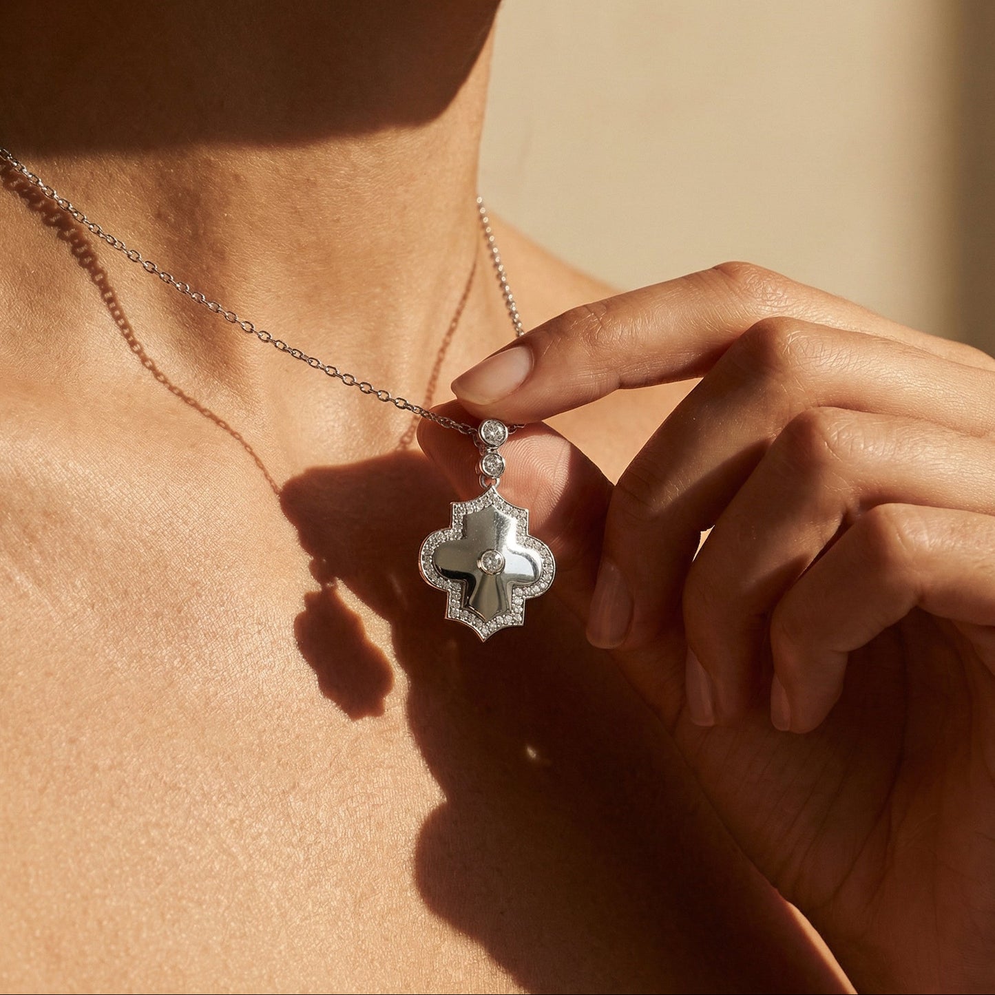 Person wearing a necklace with a decorative white gold white diamond pendant , held by a hand against a neutral background