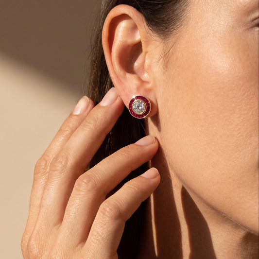 A close-up lifestyle shot of a woman wearing a round stud earring. The earring features a central clear gemstone surrounded by a double-row border of deep red gemstones, set in a yellow gold setting.