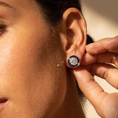 A close-up lifestyle shot of a woman wearing a small round stud earring with a dark blue outer ring and a central clear gemstone, while another hand adjusts or touches the earring.