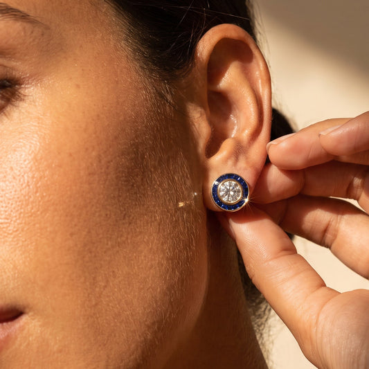 A close-up lifestyle shot of a woman wearing a small round stud earring with a dark blue outer ring and a central clear gemstone, while another hand adjusts or touches the earring.
