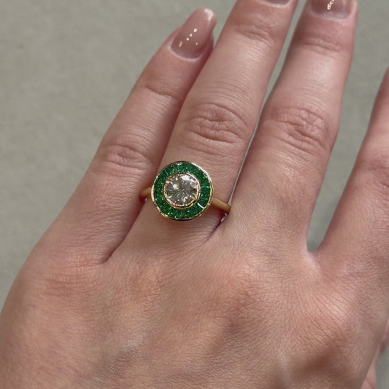 Art Deco style engagement ring on a yellow gold band with a green emerald halo and a white diamond in the center on a female hand 
