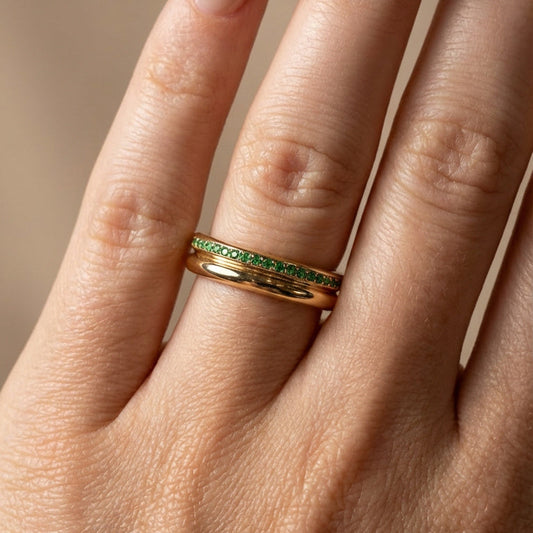 Gold ring with green gemstones on a person's finger against a neutral background